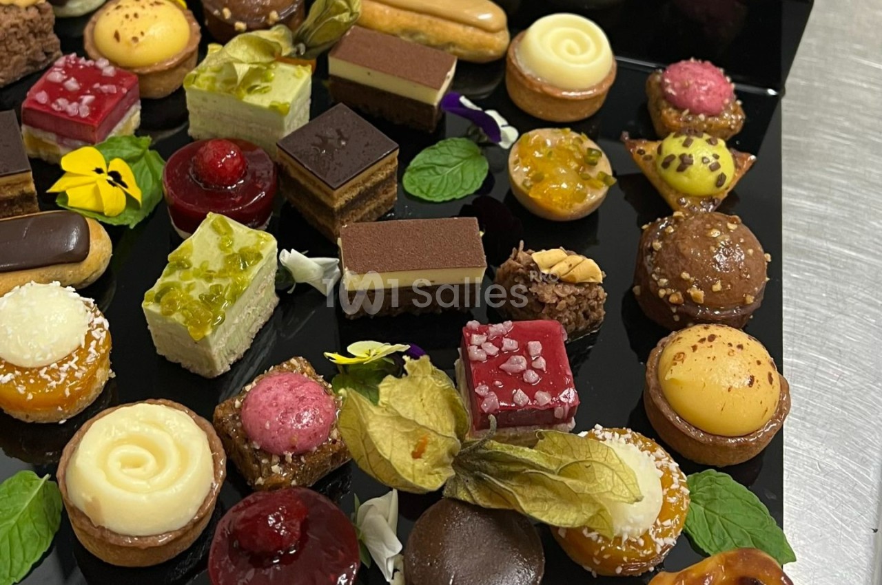 Plateau de mignardises variées comprenant tartelettes, éclairs, gâteaux et décorations florales sur fond noir.