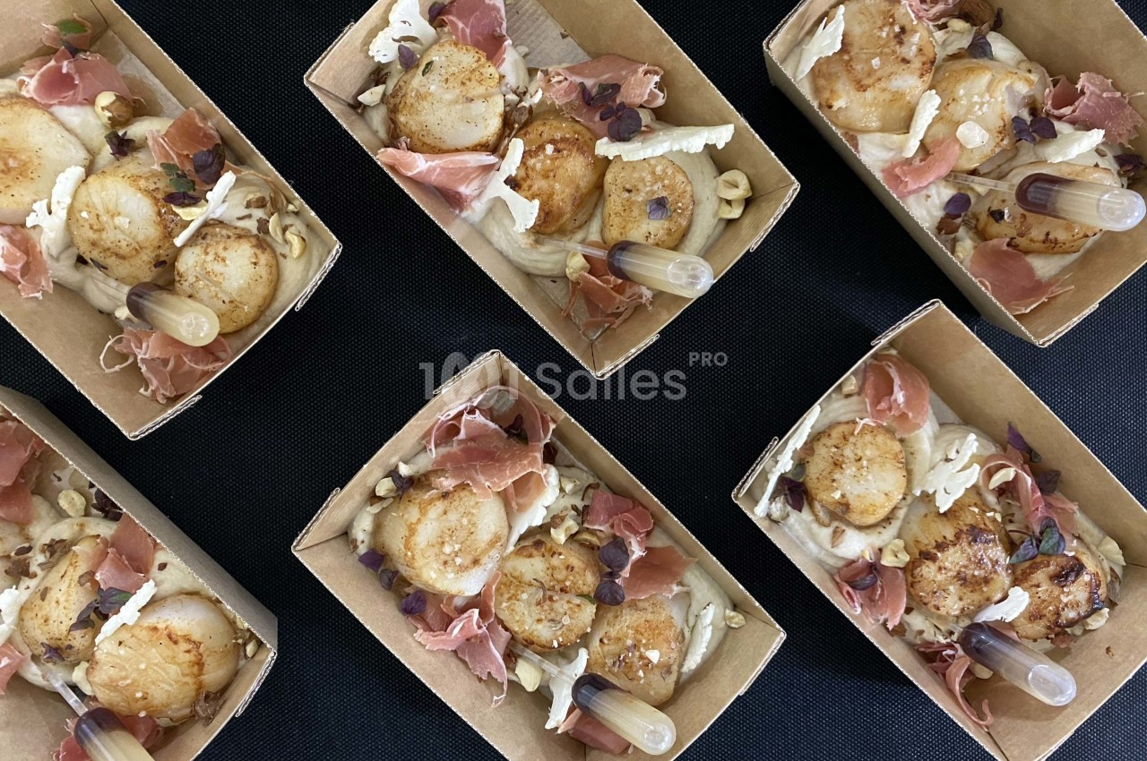 Coquilles Saint-Jacques poêlées avec jambon cru, noisettes concassées et pipettes de sauce, présentées en barquettes.