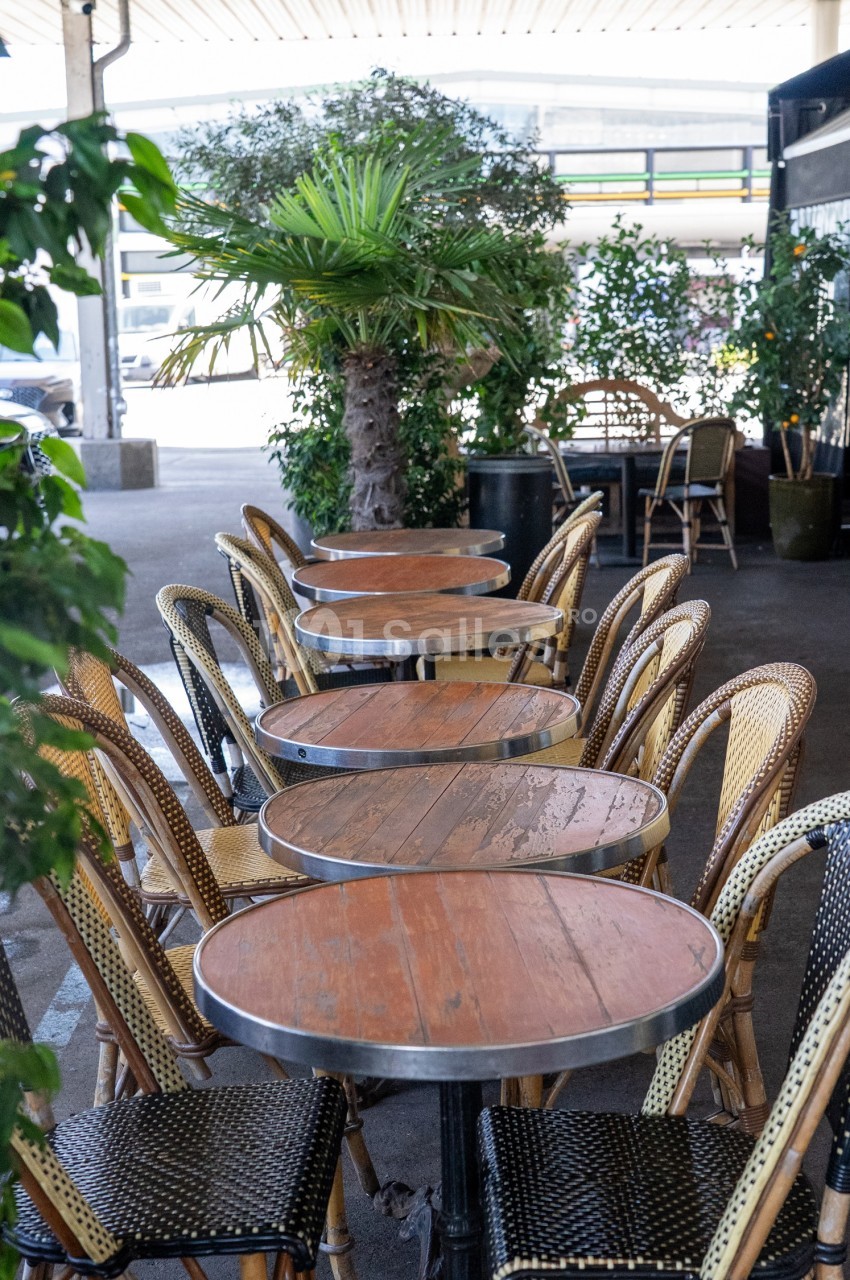 Terrasse extérieure avec des tables rondes en bois et des chaises en osier, entourées de plantes vertes.
