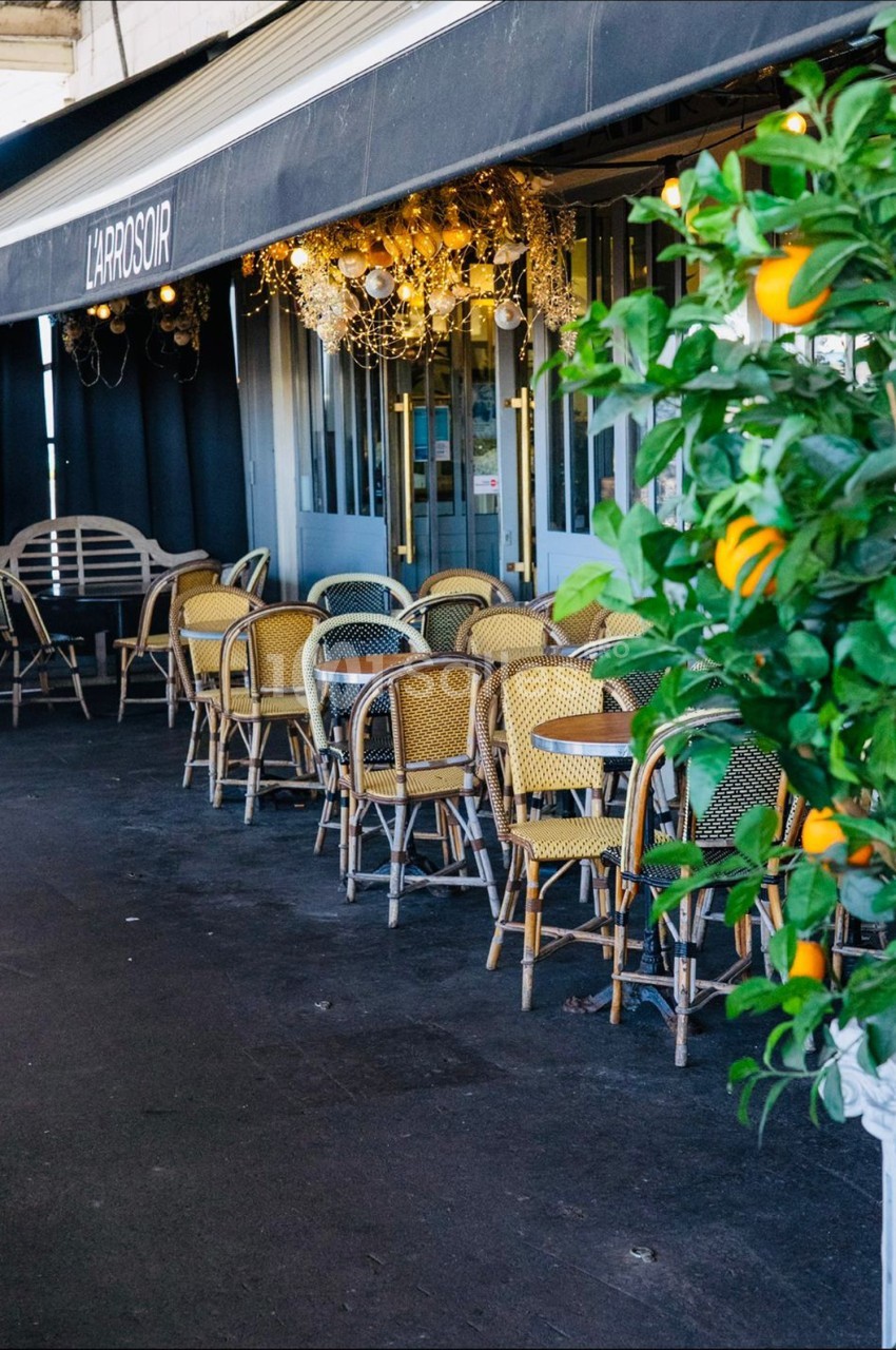 Terrasse d'un café avec des chaises en osier, une enseigne ’L'Approsoir’ et un oranger en premier plan.