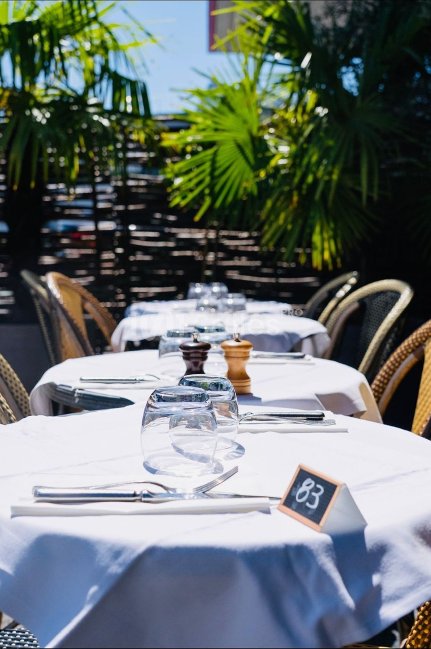 Tables de restaurant en extérieur avec nappes blanches, couverts, verres et numéro de table, entourées de plantes tropicales.