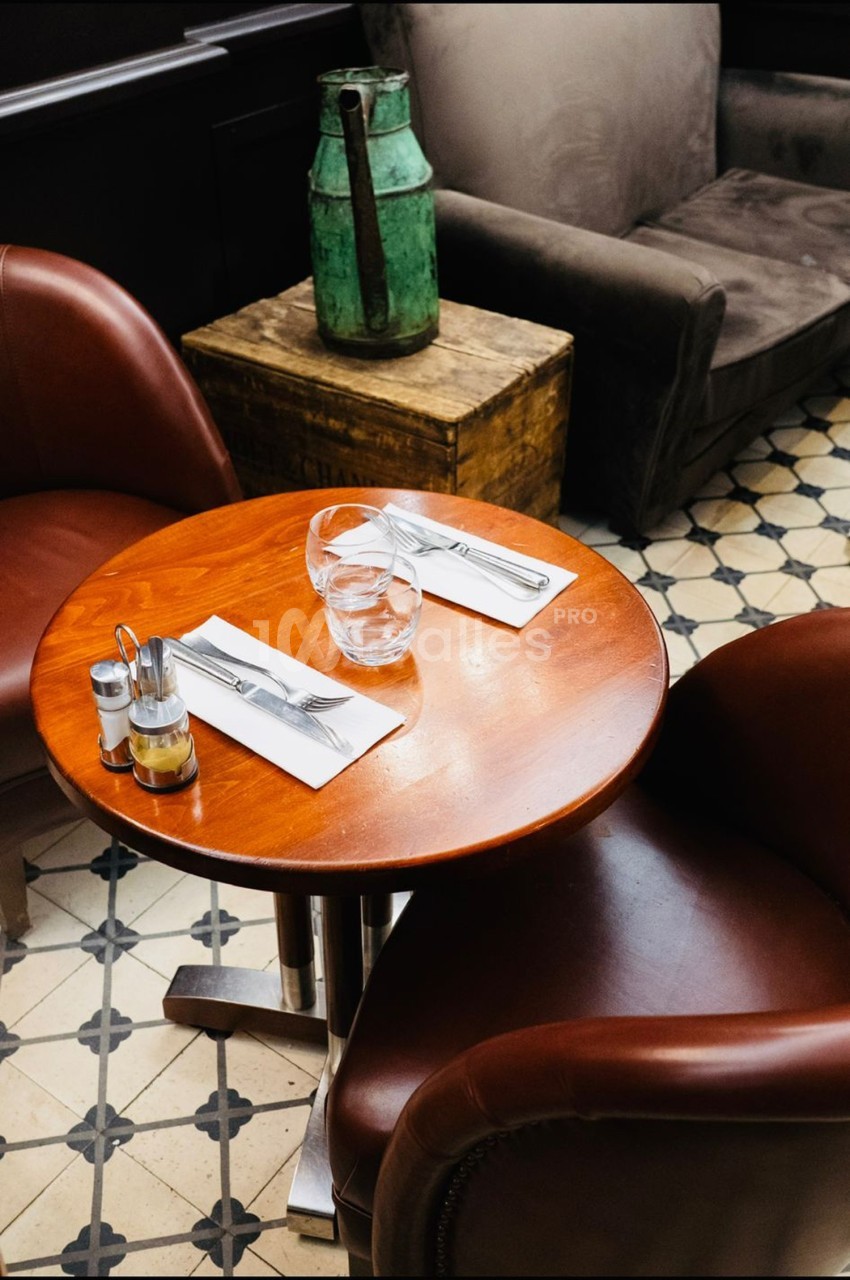 Table ronde en bois avec deux couverts, un verre d'eau et des condiments, entourée de fauteuils en cuir marron.