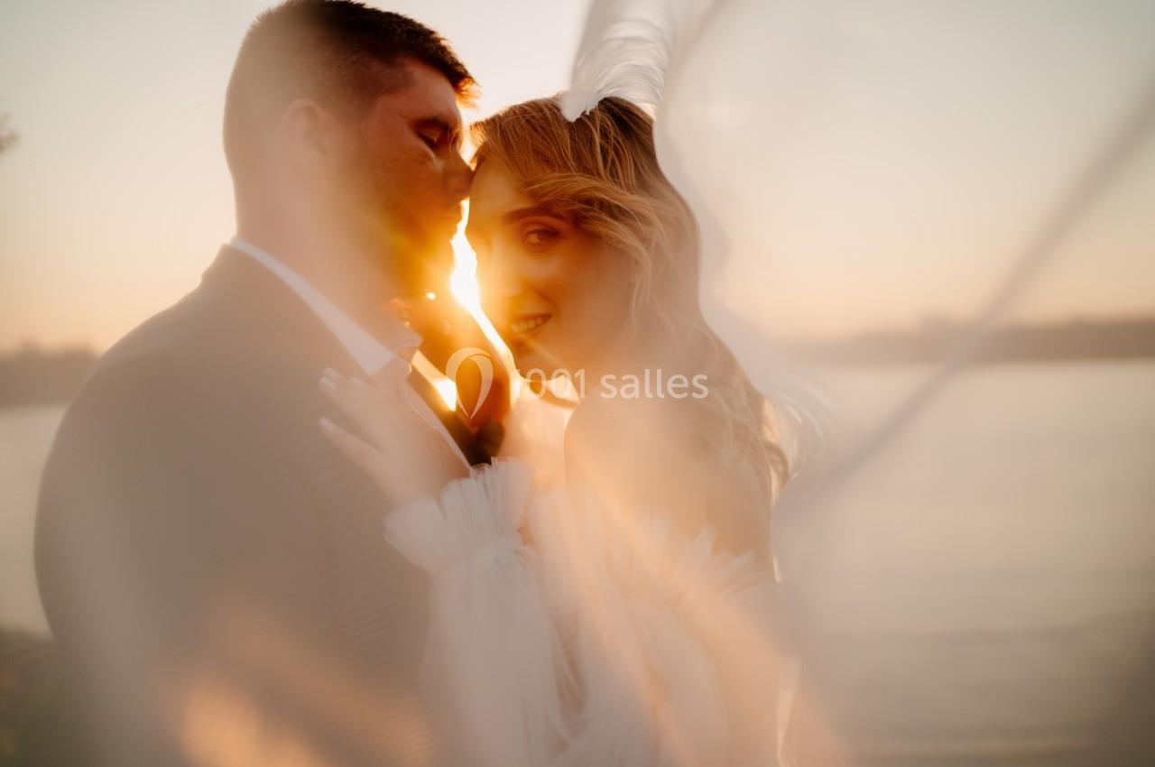 Un couple en tenue de mariage s'embrasse au coucher du soleil, avec un voile flottant devant eux.