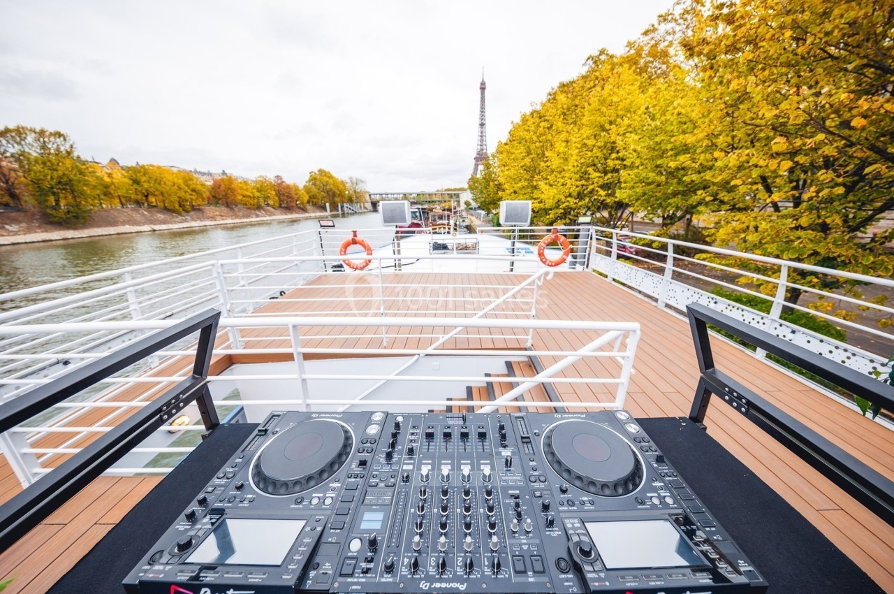 Console de DJ sur le pont d'un bateau, avec vue sur la Seine et la tour Eiffel entourée d'arbres en automne.