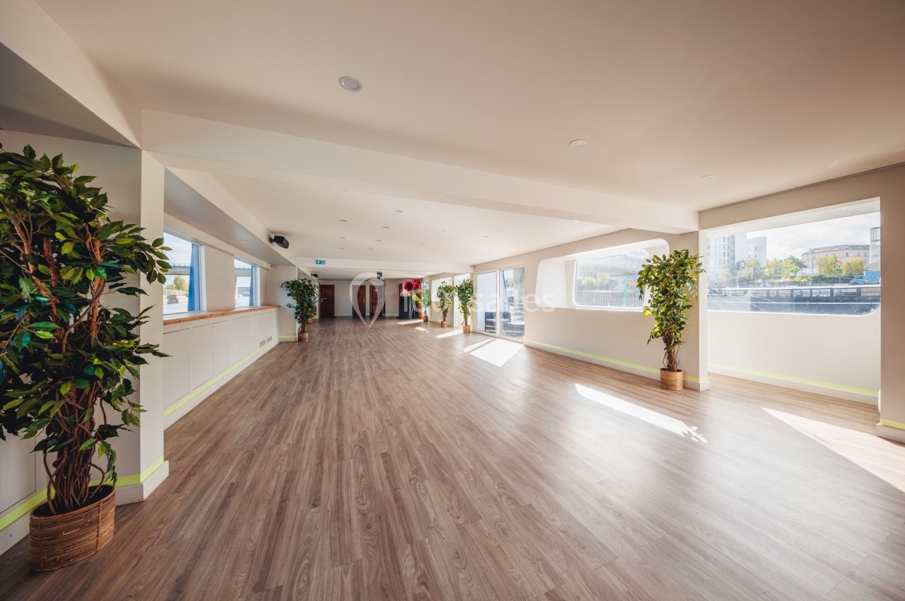 Salle lumineuse avec parquet clair, grandes fenêtres, plantes en pot et vue sur des bâtiments extérieurs.
