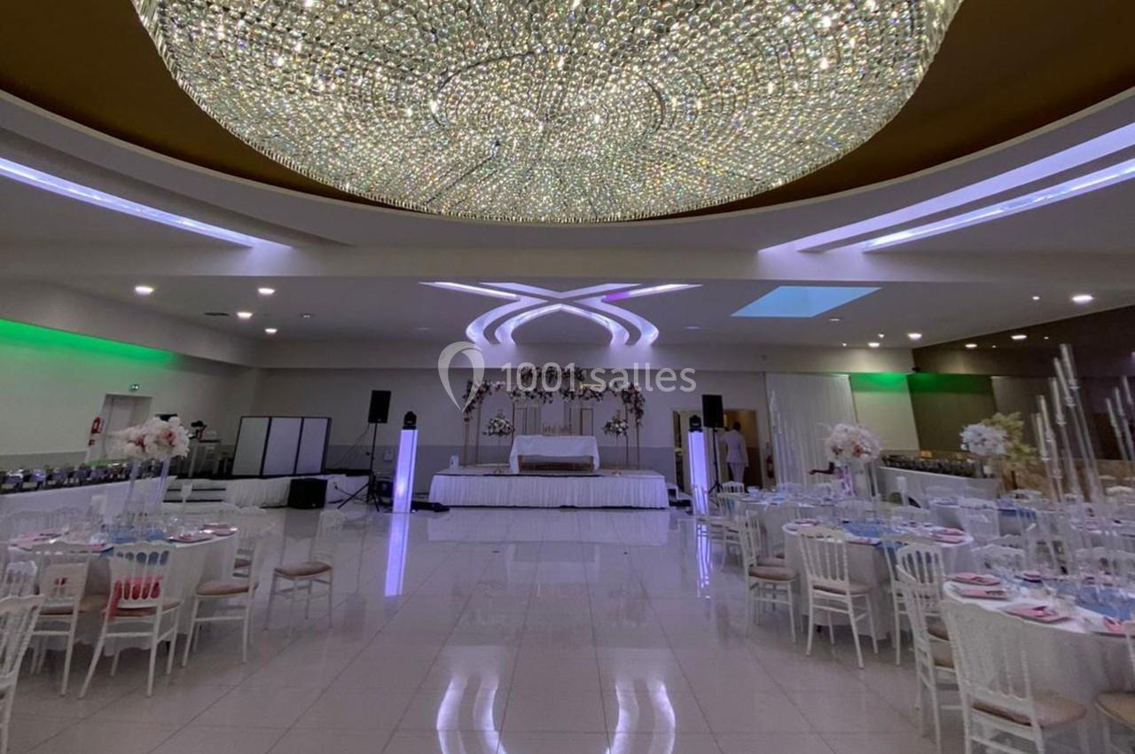 Salle de réception élégante avec tables dressées, grand lustre central et scène décorée pour un événement.