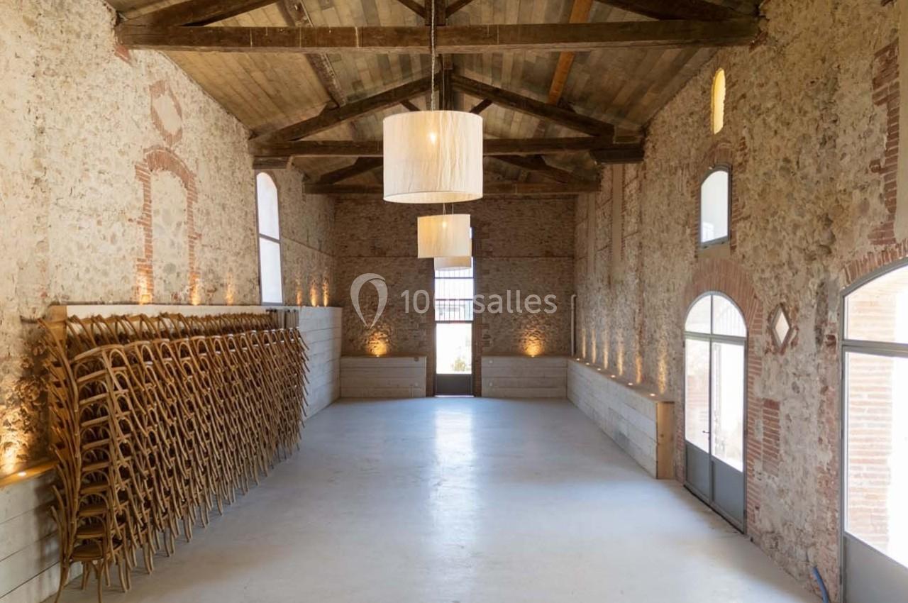 Salle lumineuse avec murs en pierre, poutres apparentes, chaises empilées sur le côté et grandes fenêtres.