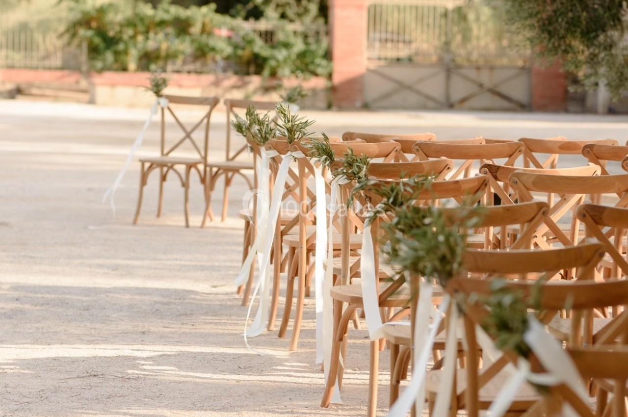 Chaises en bois alignées avec des rubans blancs et des branches d'olivier, disposées en extérieur pour un événement.