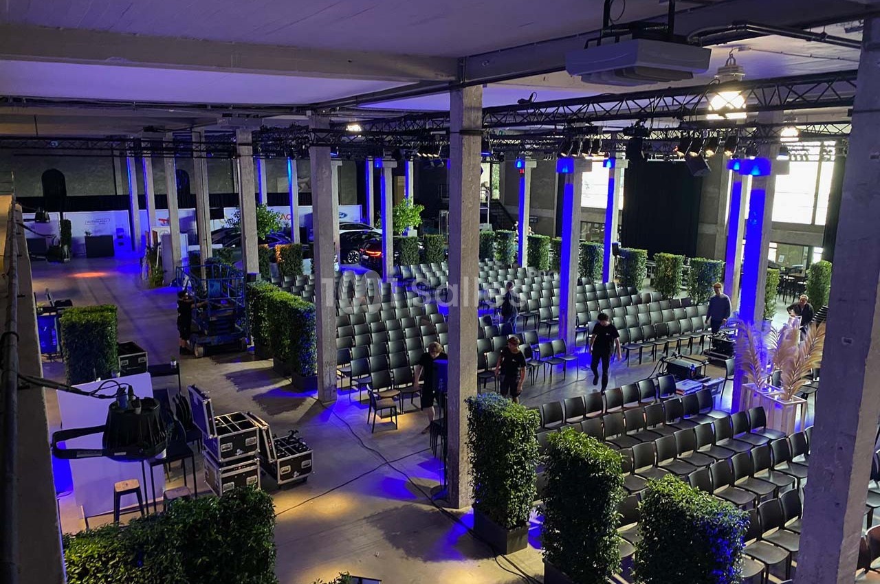 Salle de conférence avec rangées de chaises, éclairage bleu, équipements techniques et végétation décorative.