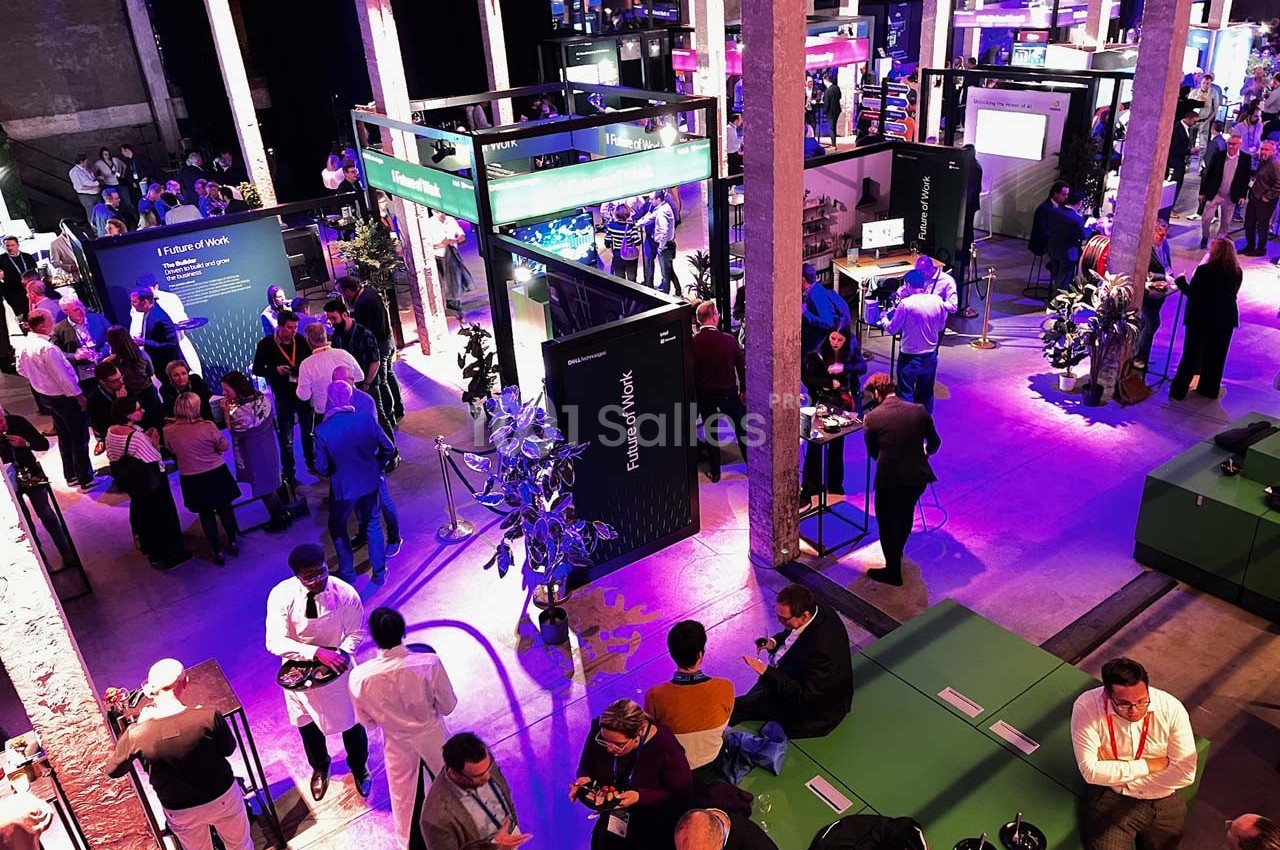Vue d'un événement professionnel en intérieur avec des stands éclairés, des participants et une ambiance animée.