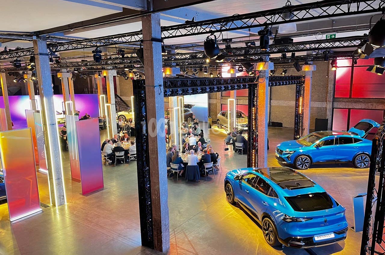 Salle d'exposition avec des voitures électriques bleues et des invités assis à des tables sous un éclairage coloré.