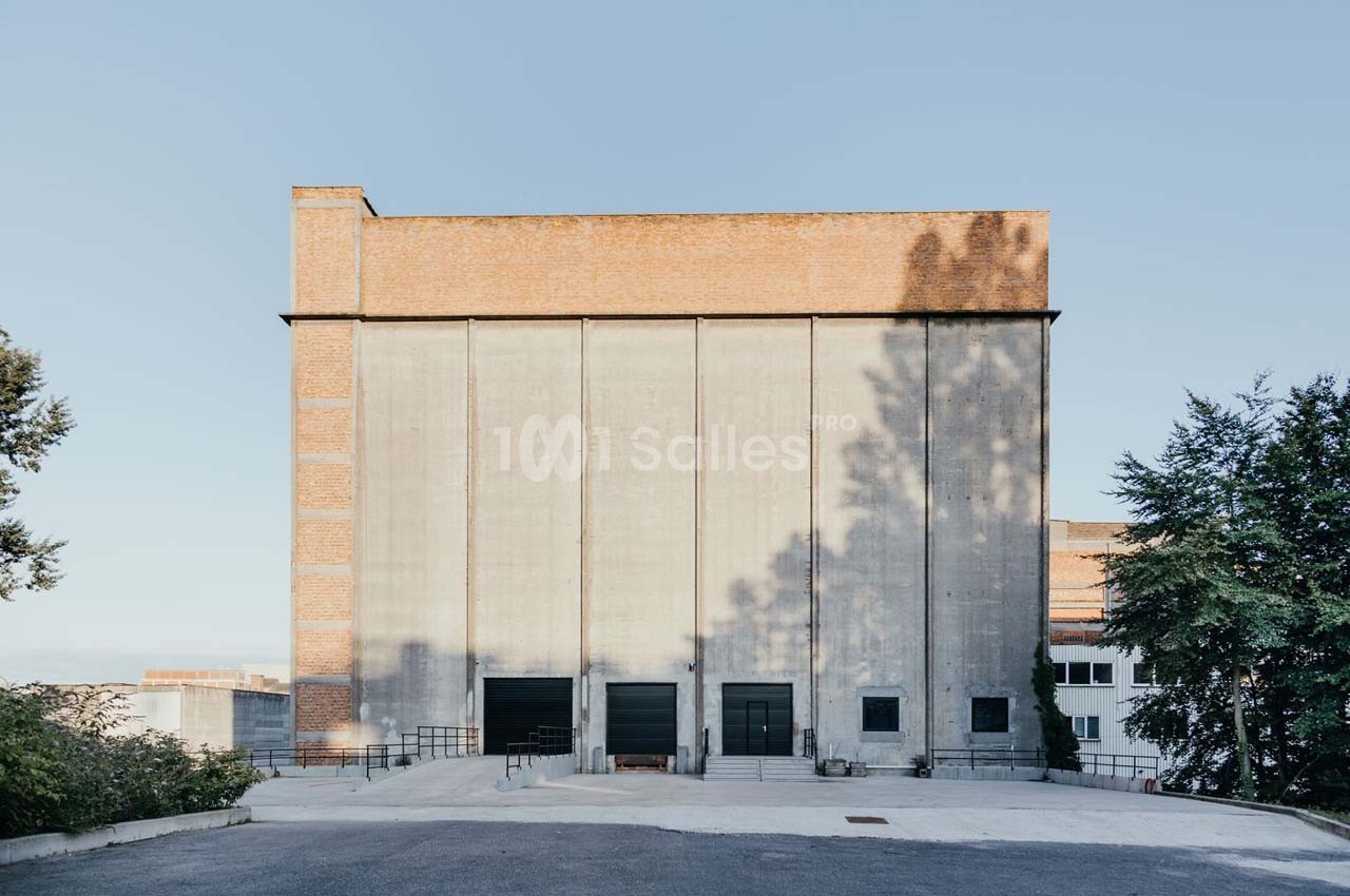 Bâtiment industriel en briques et béton avec trois grandes portes noires, entouré d'arbres et d'une cour pavée.