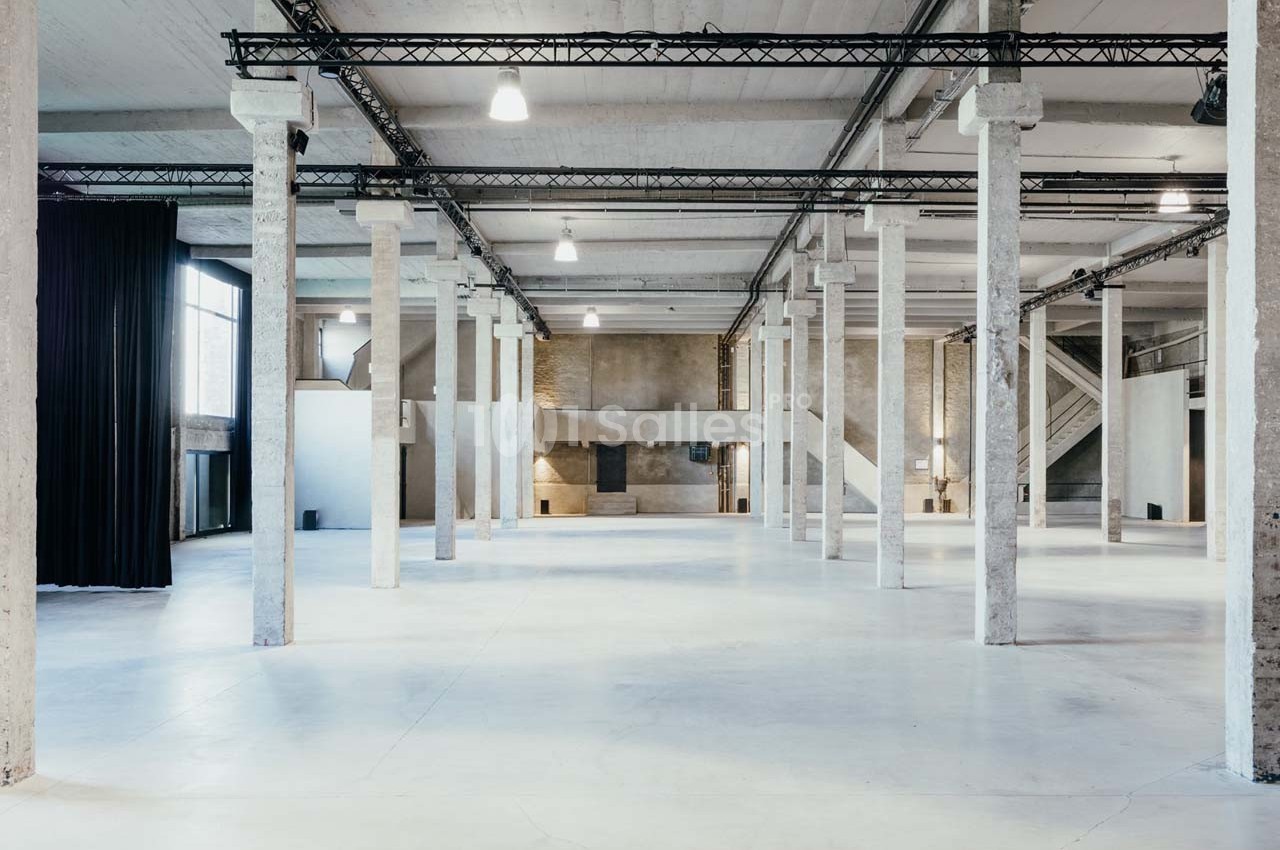 Intérieur d'un grand espace industriel lumineux avec colonnes en béton, sol clair et structures métalliques au plafond.