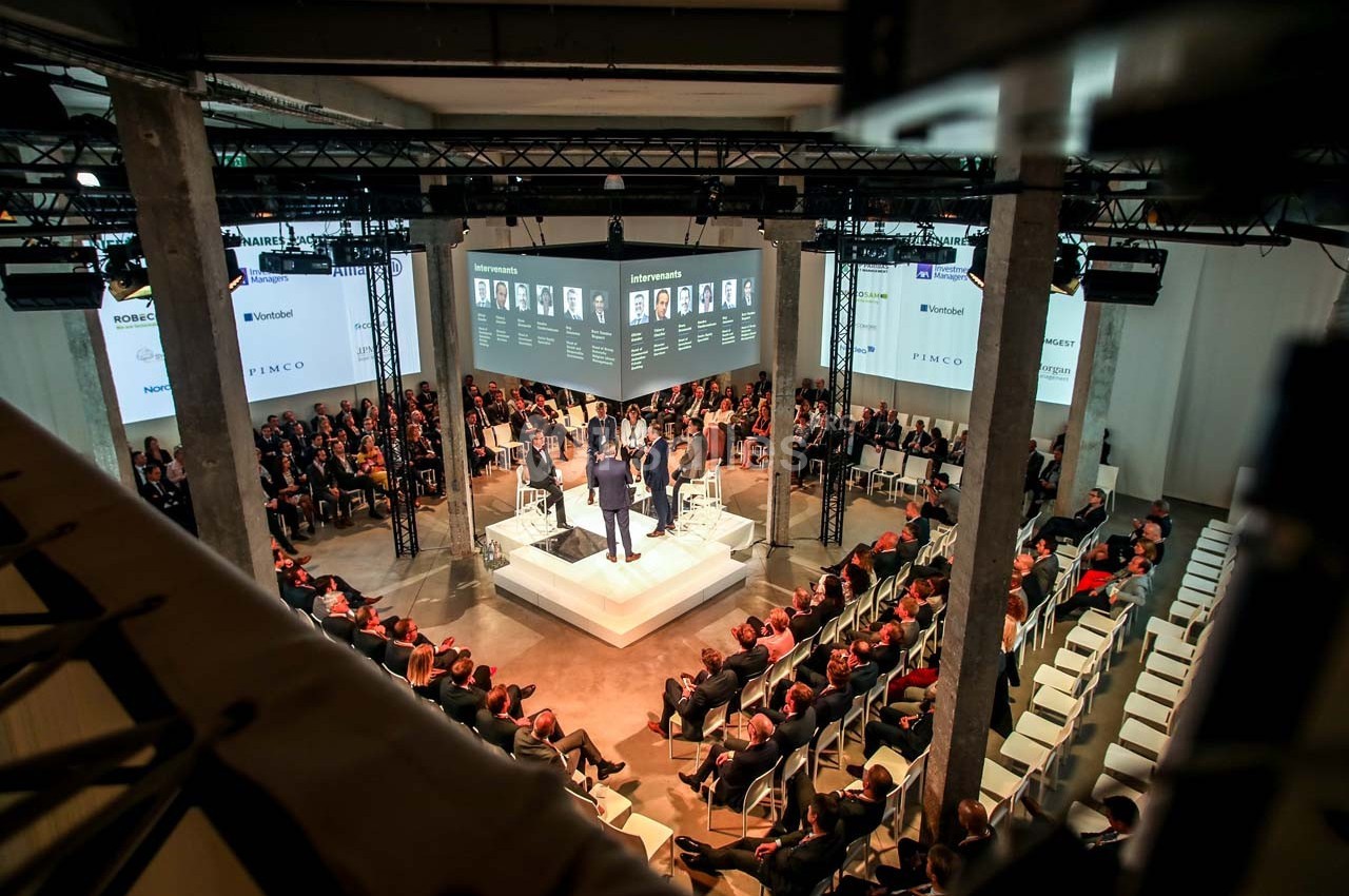 Conférence avec des intervenants sur une scène centrale, entourée d'un public assis dans une grande salle moderne.