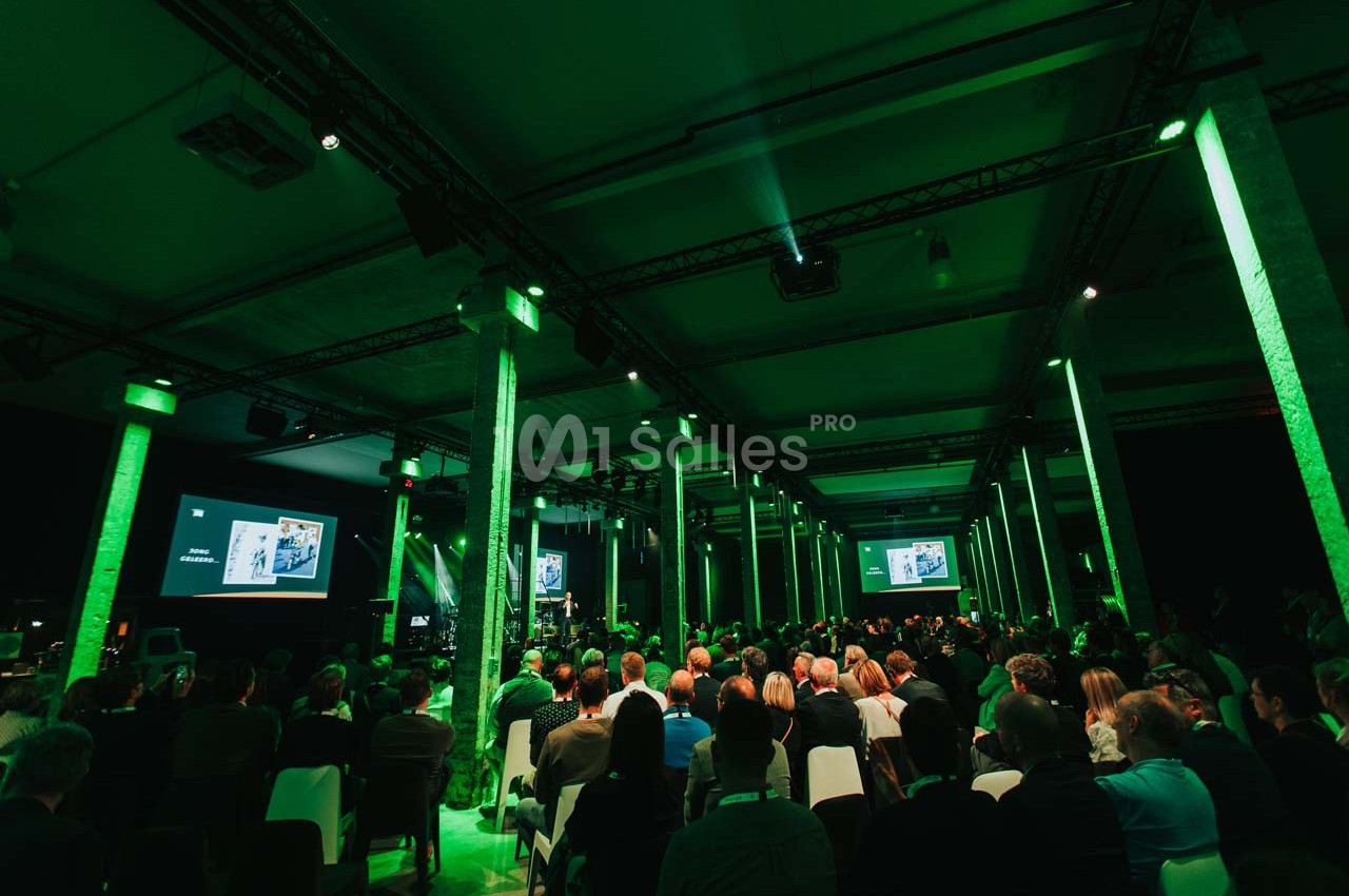 Salle de conférence avec un public assis, éclairage vert et écrans projetant des présentations.