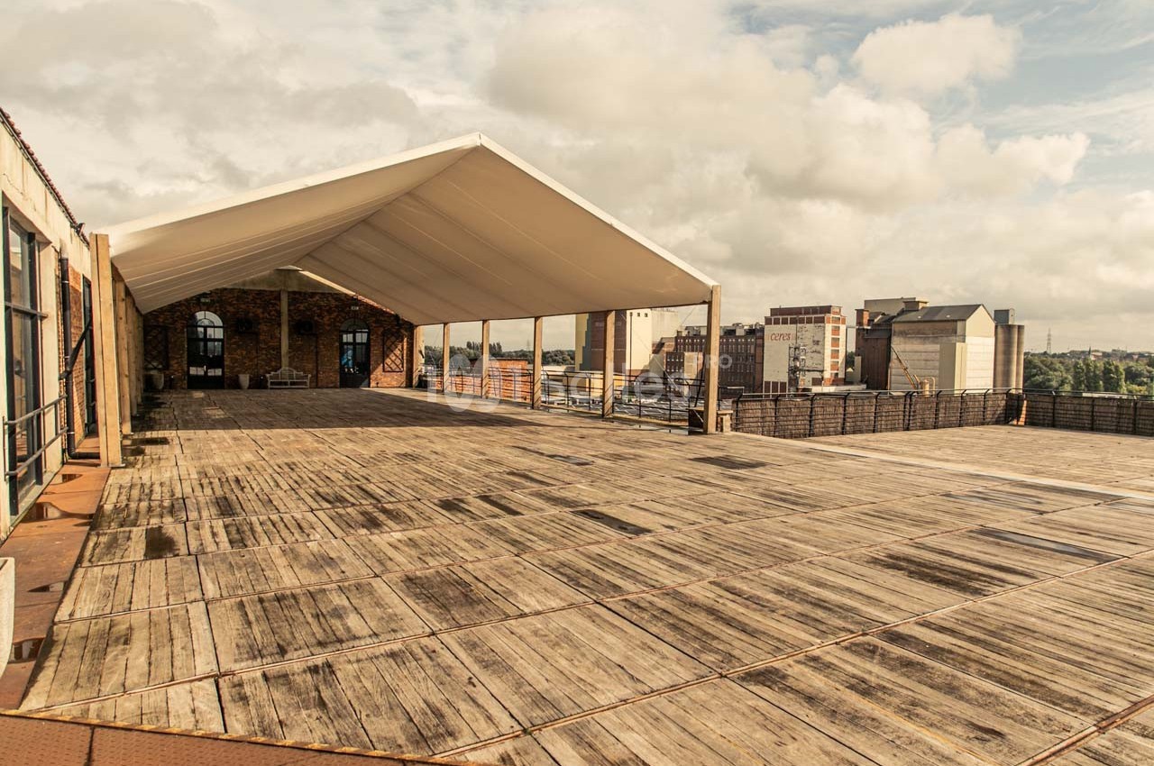 Terrasse en bois spacieuse avec une structure couverte, entourée de bâtiments urbains sous un ciel partiellement nuageux.