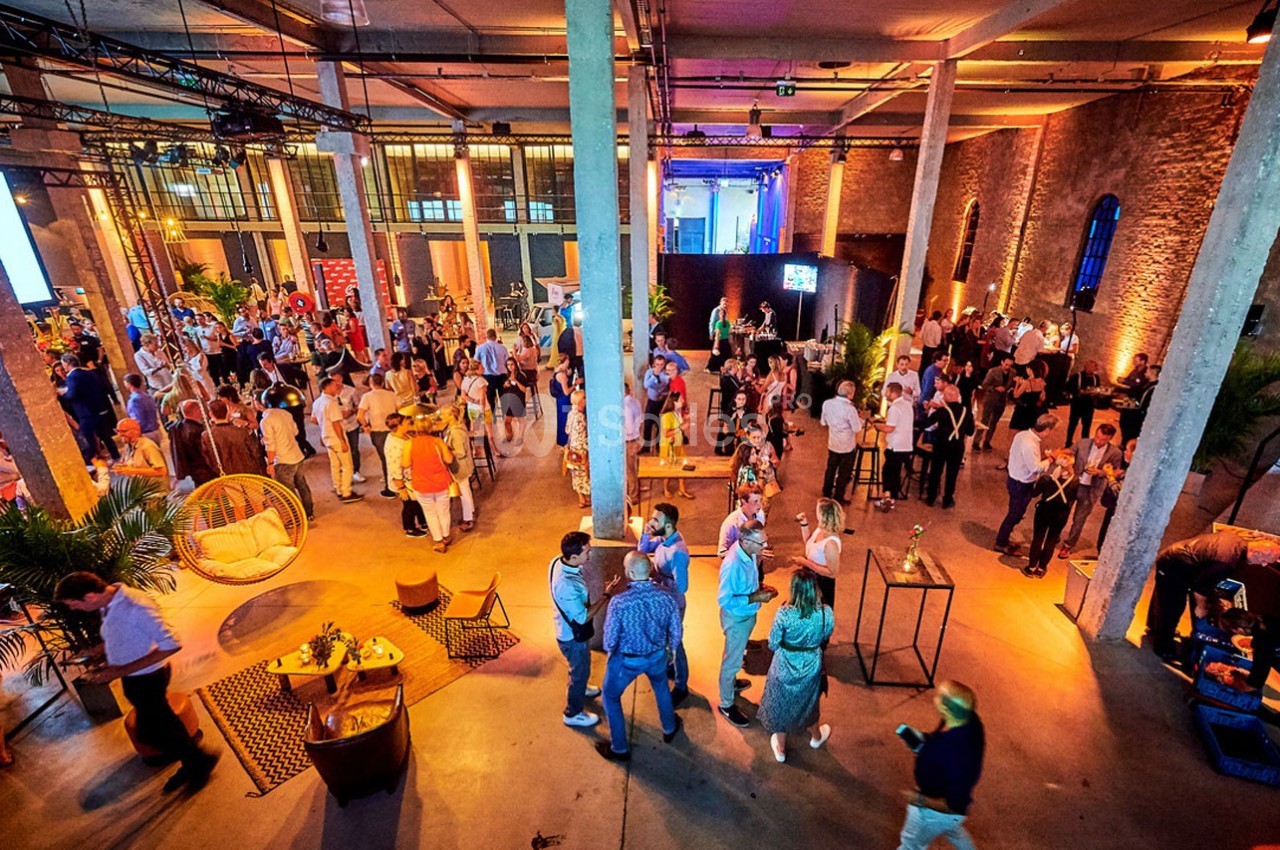 Salle spacieuse avec des invités debout et discutant lors d'un événement, éclairage chaleureux et ambiance conviviale.