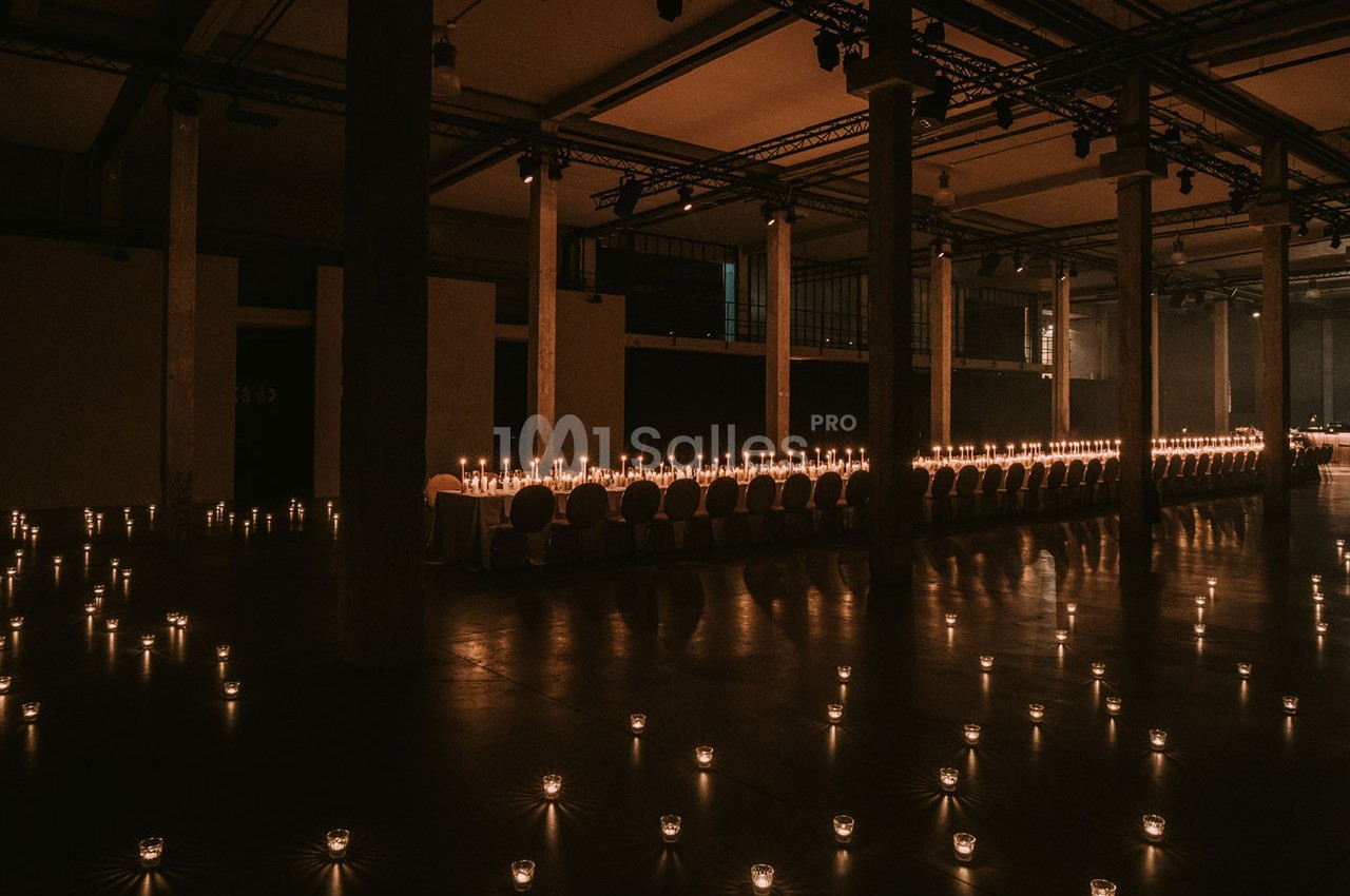 Grande salle sombre éclairée par des rangées de bougies sur une longue table et au sol, créant une ambiance chaleureuse.
