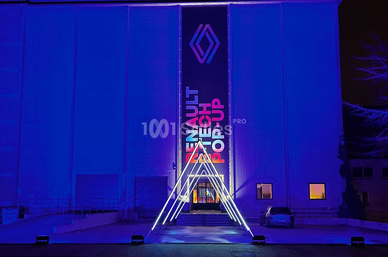 Façade d'un bâtiment illuminé en bleu avec un logo et une bannière ’Renault Tech Pop-Up’ au centre.