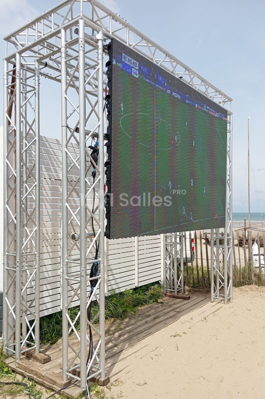 Écran géant installé sur une plage, diffusant un match de football, entouré de structures métalliques.