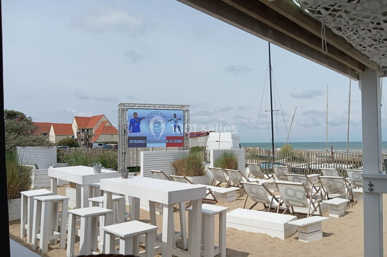 Terrasse en bord de mer avec tables et chaises blanches, vue sur la plage, un écran et des voiliers en arrière-plan.