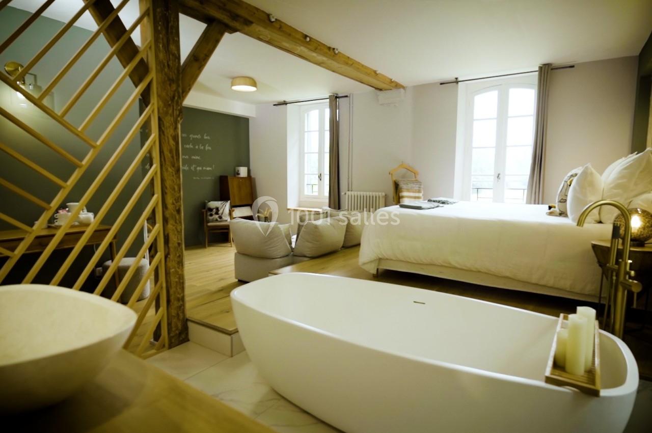 Chambre lumineuse avec baignoire îlot, lit double, poutres apparentes et grandes fenêtres donnant sur l'extérieur.