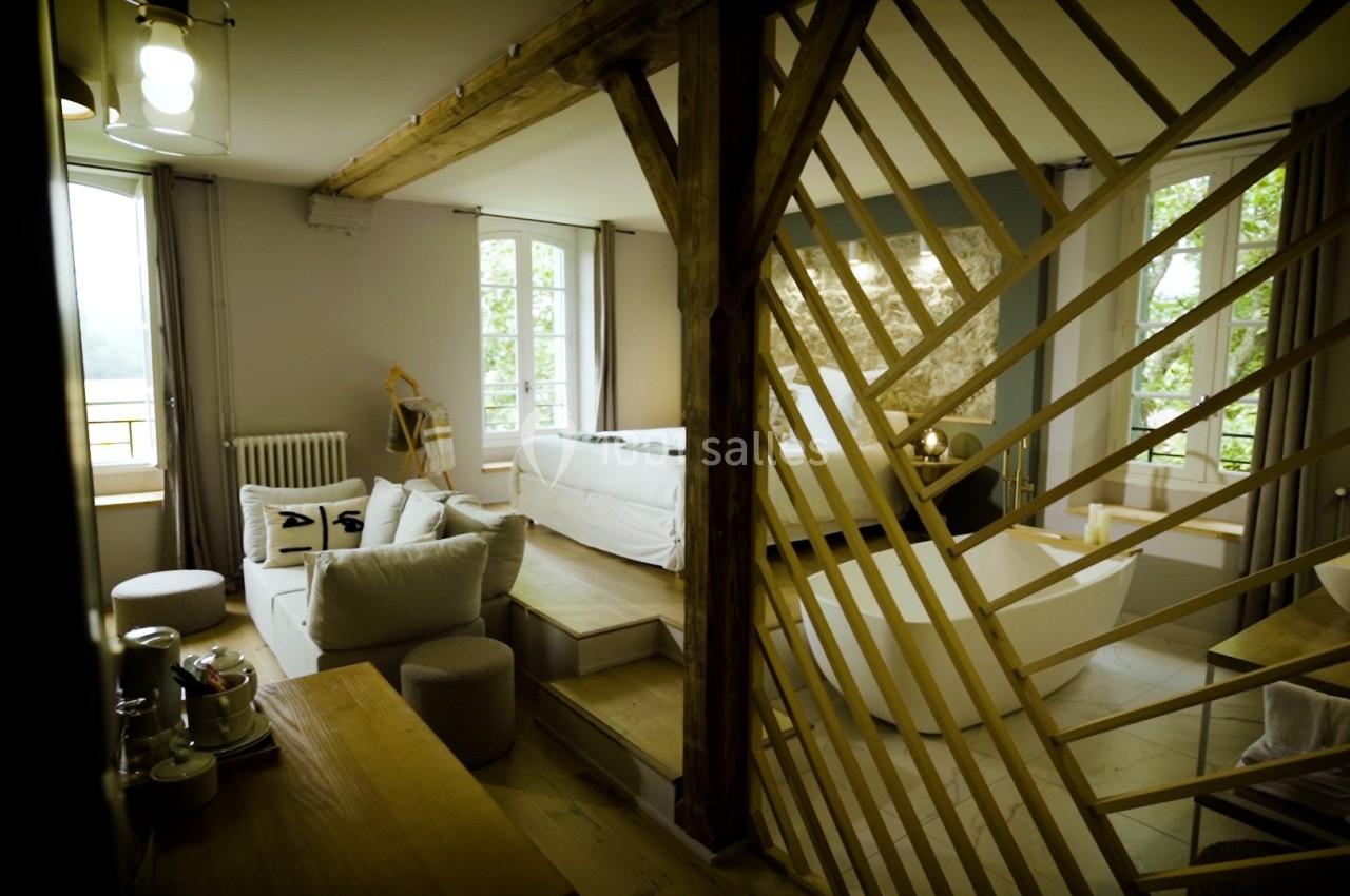 Intérieur lumineux avec poutres apparentes, cloison en bois, canapé blanc et grandes fenêtres donnant sur l'extérieur.