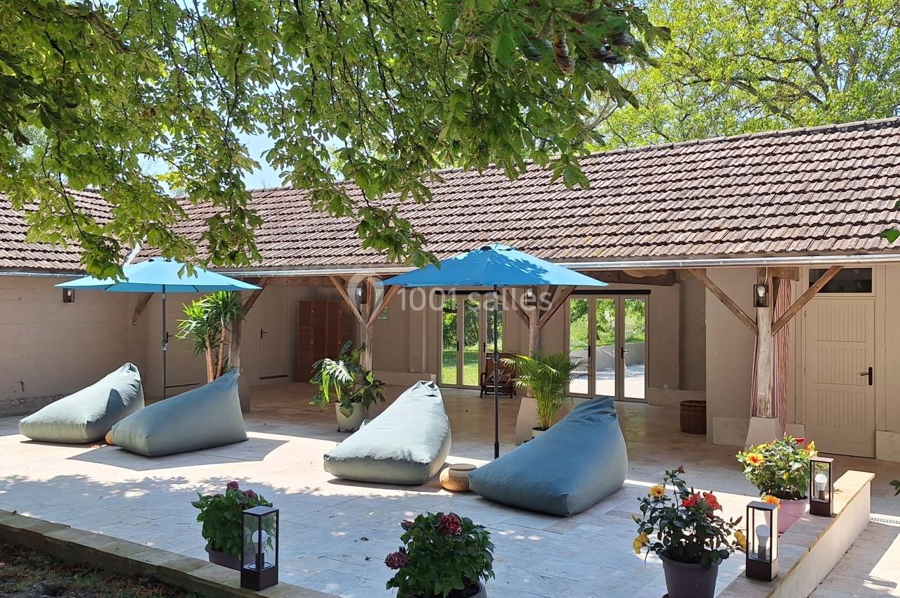Terrasse ensoleillée avec coussins de sol, parasols bleus, plantes en pot et vue sur une maison en arrière-plan.