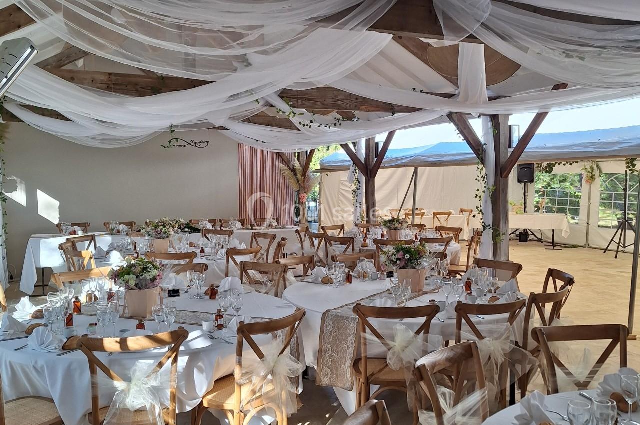 Salle de réception décorée avec des tables rondes, nappes blanches, chaises en bois et voilages suspendus au plafond.