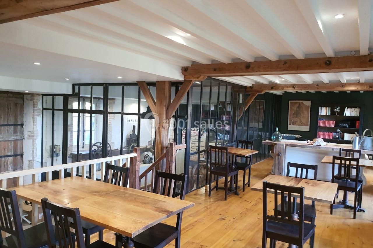 Salle lumineuse avec tables en bois, chaises noires, poutres apparentes et verrières donnant sur un espace adjacent.