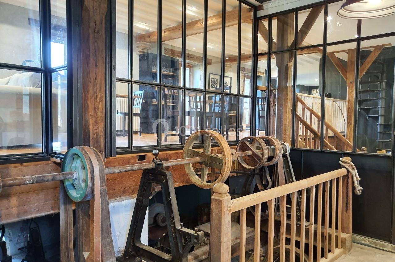 Mécanisme ancien en bois et métal devant une verrière, dans un espace intérieur lumineux avec poutres apparentes.