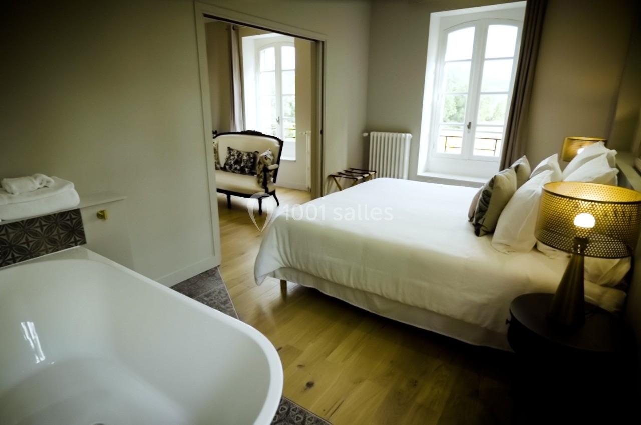 Chambre lumineuse avec lit double, baignoire blanche au premier plan et fauteuils près des fenêtres.