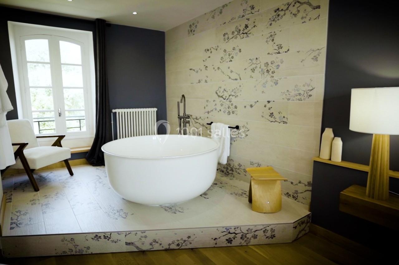 Salle de bain élégante avec baignoire ronde, mur décoré de motifs floraux, fauteuil et lampe en bois clair.