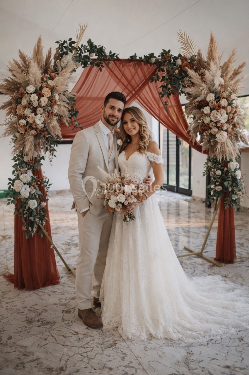 Un couple en tenue de mariage pose devant une arche décorée de fleurs et de drapés rouges.