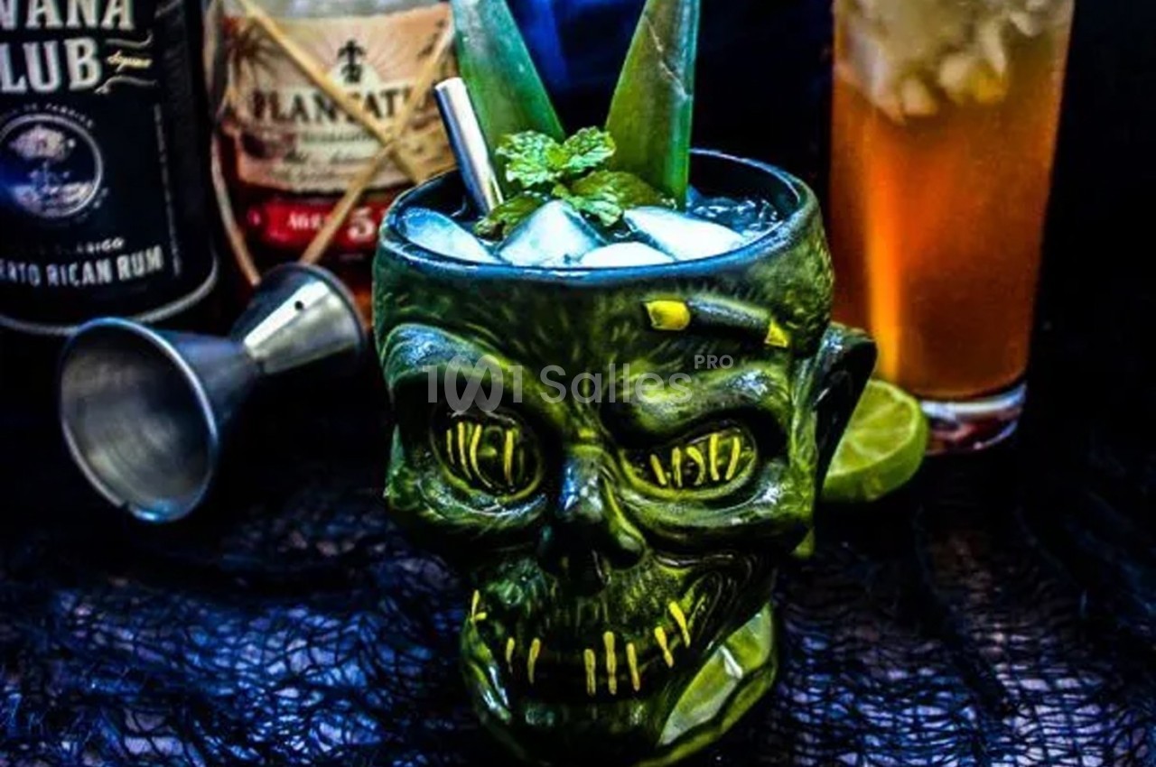 Verre en forme de tête de zombie rempli d'un cocktail glacé, décoré de feuilles et de menthe, avec des bouteilles en arrière…