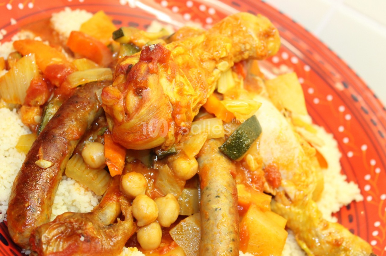 Plat de couscous avec légumes, pois chiches, merguez et pilon de poulet, servi sur une assiette rouge.