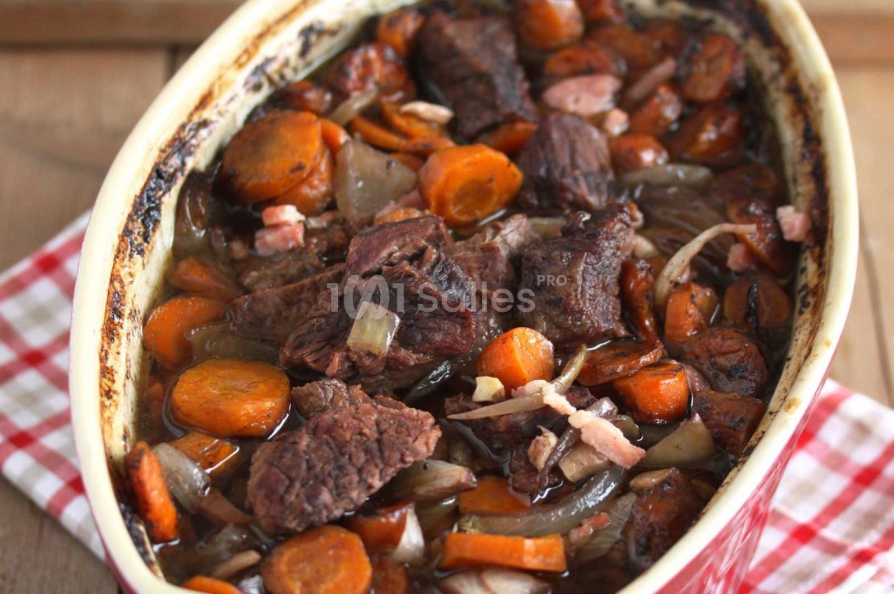 Plat de bœuf mijoté avec des carottes, oignons et lardons dans un plat en céramique sur une nappe à carreaux rouges.