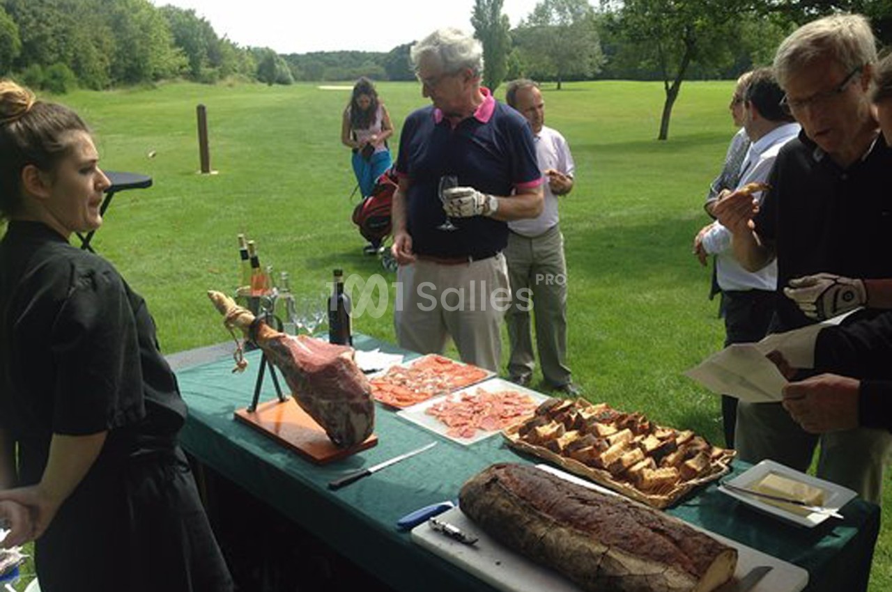 Des personnes dégustent des plats à base de charcuterie et de pain autour d'une table en plein air sur un terrain verdoyant.