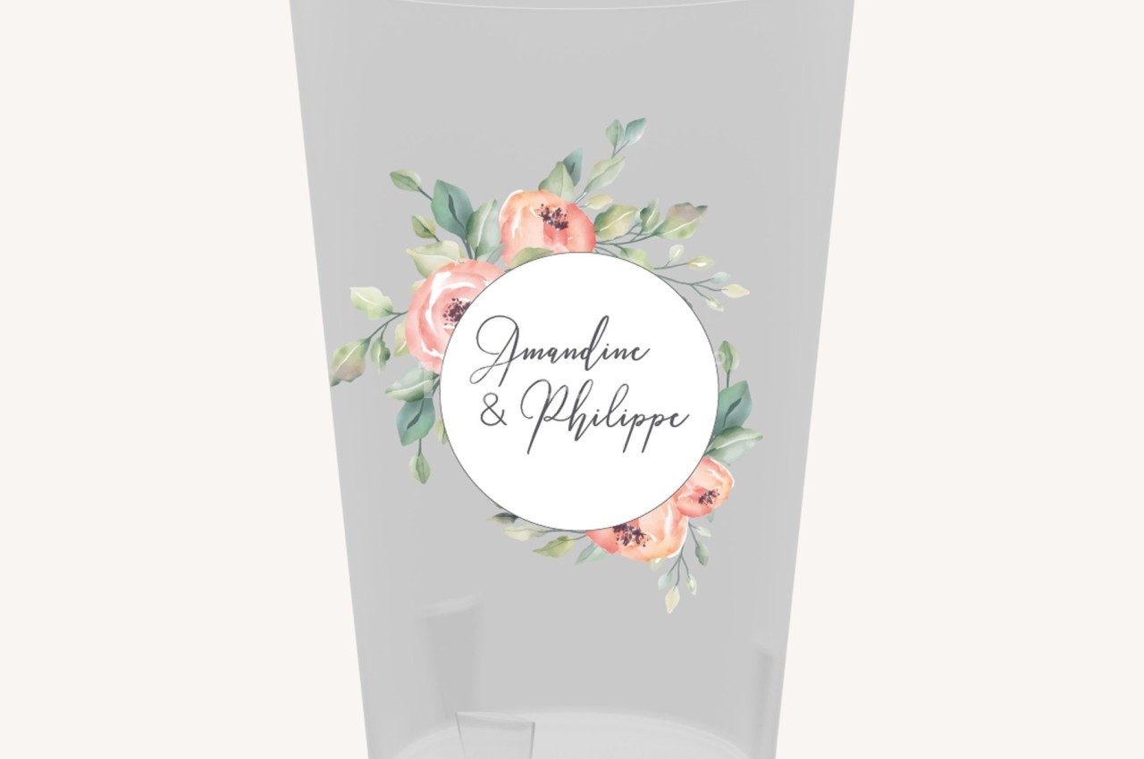 Verre transparent décoré d'une étiquette ronde avec des fleurs pastel et des prénoms manuscrits au centre.