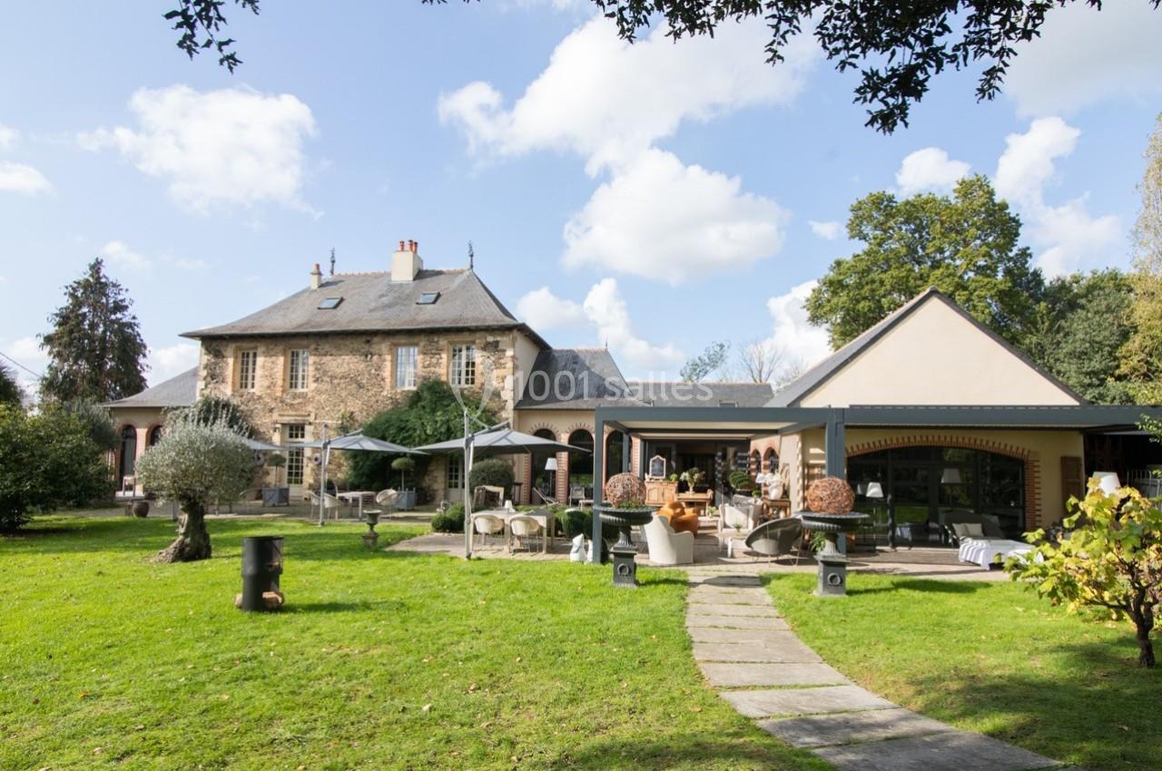 Grande maison en pierre avec extension moderne, entourée d'un jardin verdoyant sous un ciel partiellement nuageux.