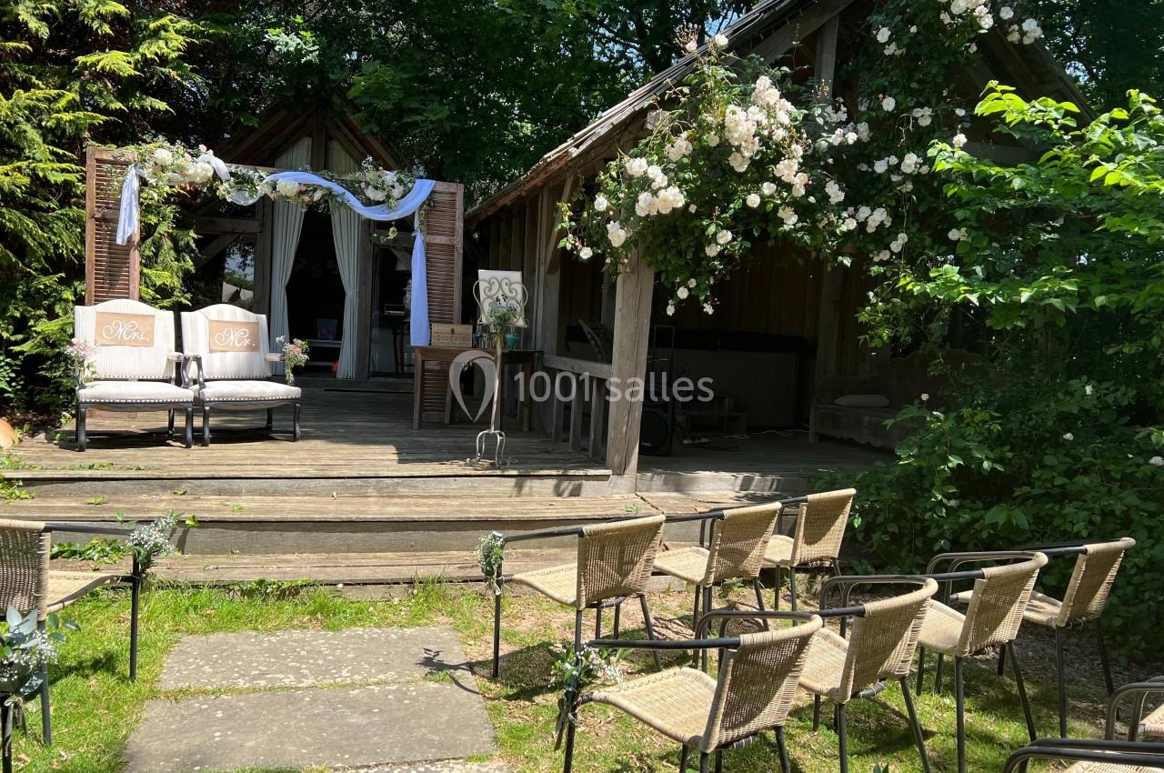 Chaises en osier disposées devant une petite scène en bois décorée de fleurs blanches et de voilages, en extérieur.