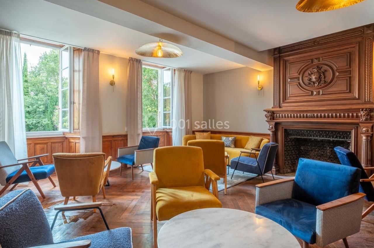 Salle lumineuse avec parquet, cheminée en bois sculpté, fauteuils colorés et grandes fenêtres ouvertes sur un jardin.