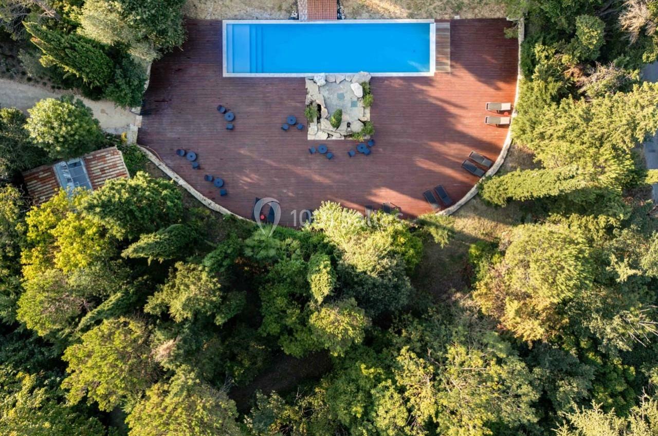 Vue aérienne d'une piscine rectangulaire entourée d'une terrasse en bois avec chaises, au milieu d'une végétation dense.