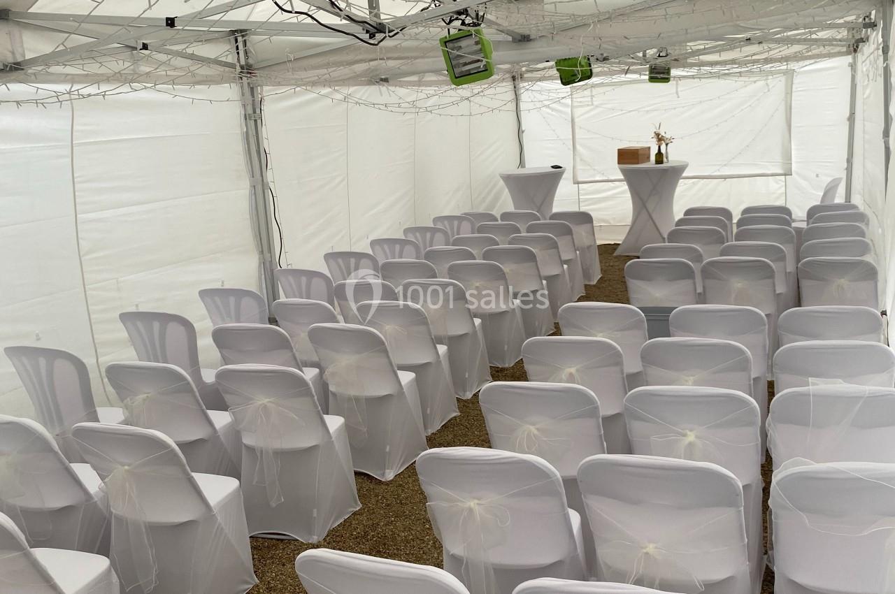 Chaises blanches alignées sous une tente blanche, disposées face à une table avec des objets décoratifs.