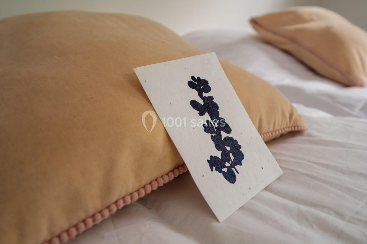 Carte blanche avec illustration florale bleue posée sur un coussin beige, sur un lit aux draps blancs.