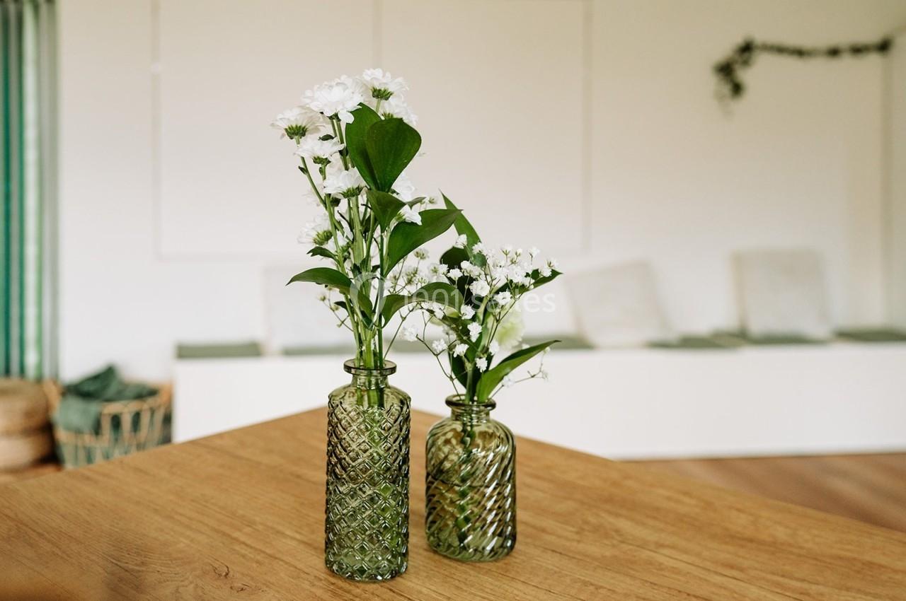 Deux vases en verre texturé contenant des fleurs blanches et des feuilles vertes, posés sur une table en bois clair.