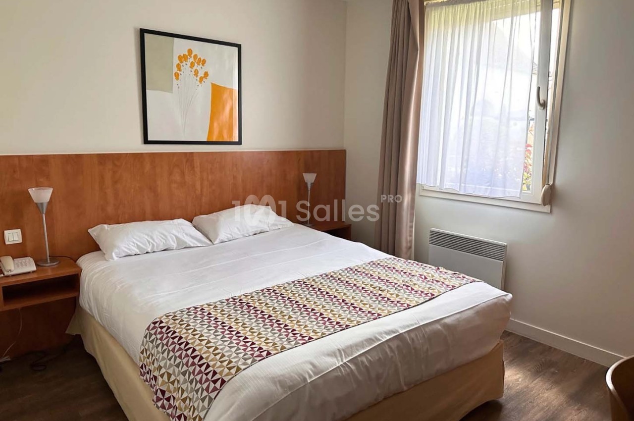 Chambre avec lit double, tête de lit en bois, tableau décoratif, lampe de chevet et fenêtre avec rideaux.