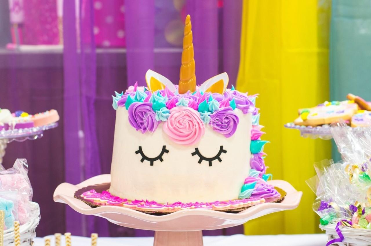 Gâteau décoré en forme de licorne avec des fleurs colorées et une corne dorée, présenté sur un présentoir rose.