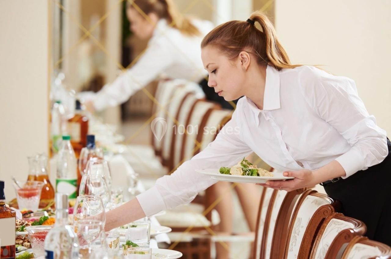 Une serveuse en chemise blanche dresse une table élégante avec des plats et des boissons dans un cadre lumineux.