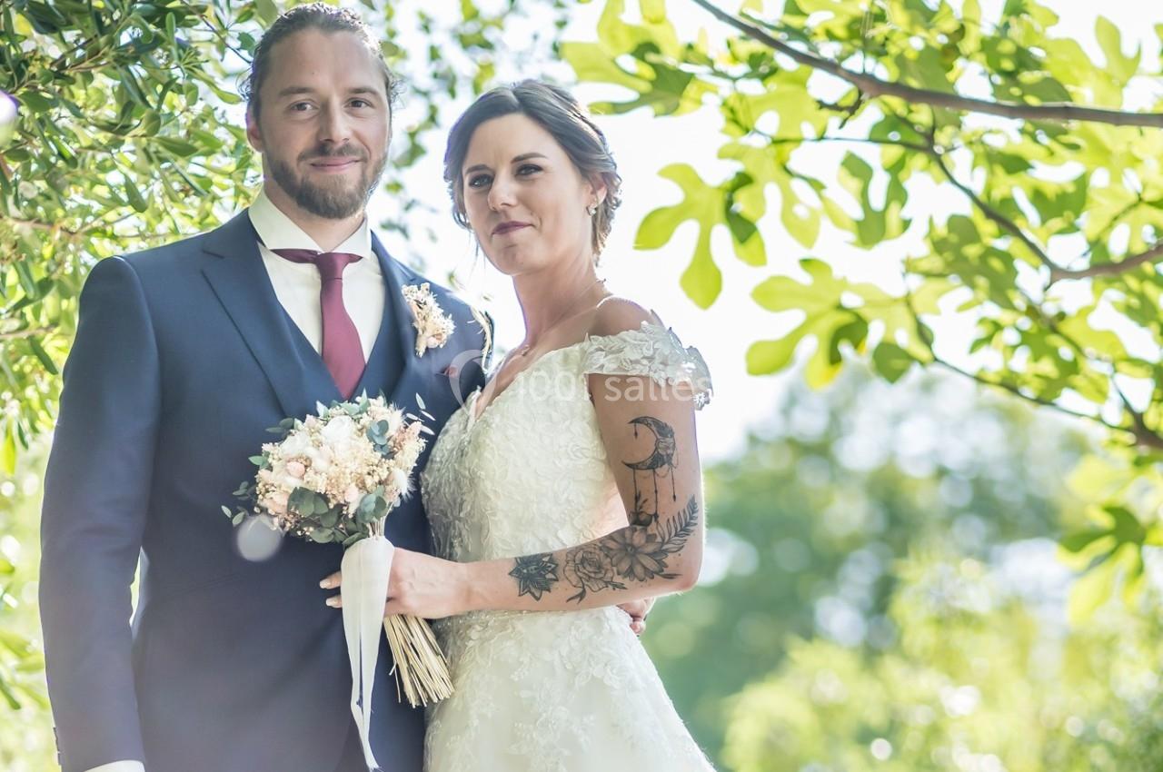 Un couple en tenue de mariage pose dans un jardin ensoleillé, entouré de feuillage vert.